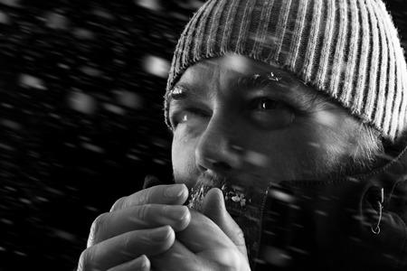 Freezing cold man standing in a snow storm biazzard trying to keep warm. Wearing a beanie hat and winter coat with frost and ice on his beard and eyebrows. Black and white.