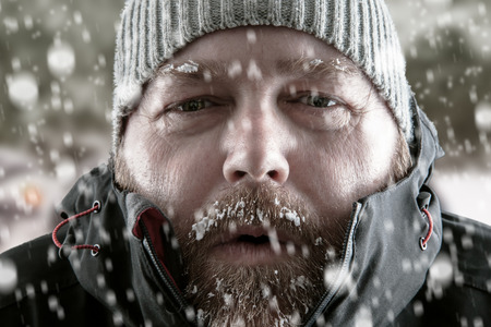 Freezing cold man standing in a snow storm blizzard trying to keep warm. Wearing a beanie hat and winter coat with frost and ice on his beard and eyebrows staring at the camera.