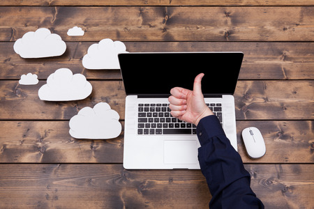 Cloud computing technology concept with white fluffy clouds next to the laptop. Man with thimbs up over the  keyboard uploading data on a wooden table.の写真素材