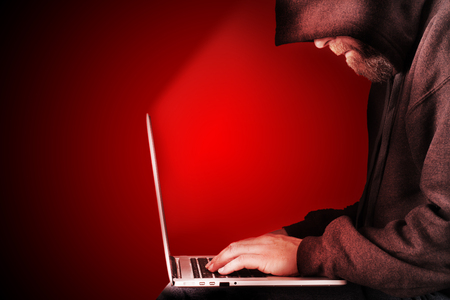 Male computer hacker wearing a hooded top leaning over a laptop with red warning background. The screen light illuminates the man with a beard performing illegal activities. Graduated copy space on the leftの写真素材