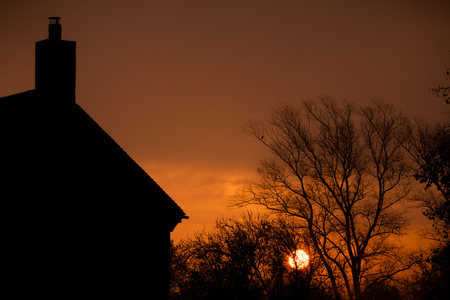 Sunrise at home. Early morning sun glowing through misty morning autumn trees.  Relaxing rural landscape at dawn with an orange sun rising behind a silhouette of a house and bare trees.の写真素材