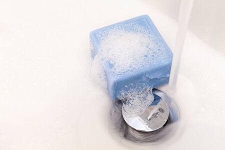 Bar of square blue soap and suds in a white sink with chrome plug and runnning water tap. Copy spaceの写真素材