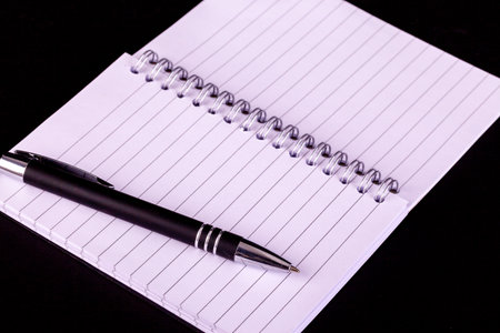 Pen and opened A5 notepad with white lined pages and spiral boound with a black backgroundの写真素材
