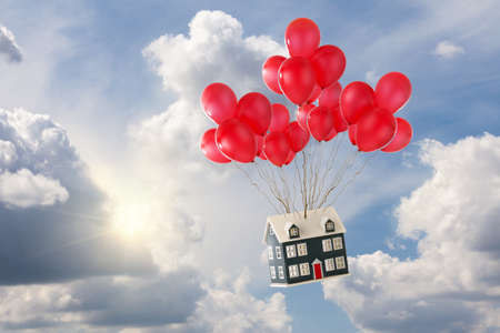 Toy house with red balloons floating in the blue sky with sunshine and white fluffy clouds. Soaring house princes and moving home conceptの写真素材