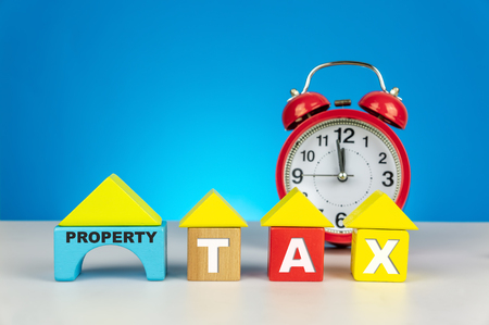 Property Tax alert. Clock with house wood cube on blue background. Tax and taxation concept.の写真素材