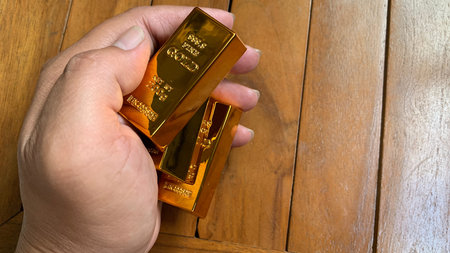 Hand holding multiple gold bars, highlighting size, weight impression, and polished surface against wooden table background