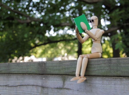 Wooden artist manikin sitting on a wooden fence and reading a green book の写真素材