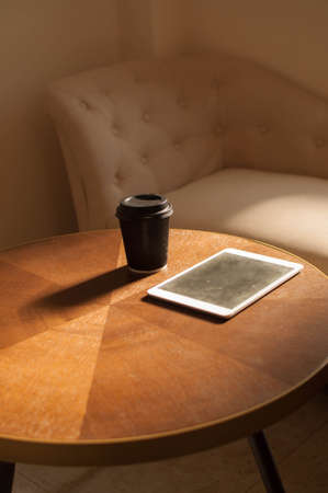 A disposable cup of coffee and a digital tablet on a wooden table indoor with sunbeamの写真素材