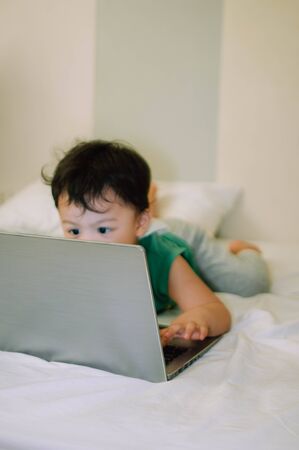 A portrait of a male toddler is playing with a laptop while on the bed