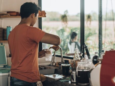 A man wearing an orange shirt standing poured coffee into a glass with ice near the window.の写真素材