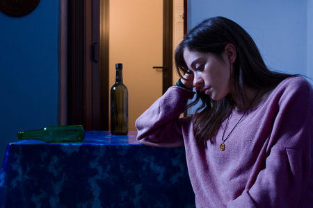 Sad Woman Sitting at the Table with Empty Alcohol Bottles at Nightの写真素材