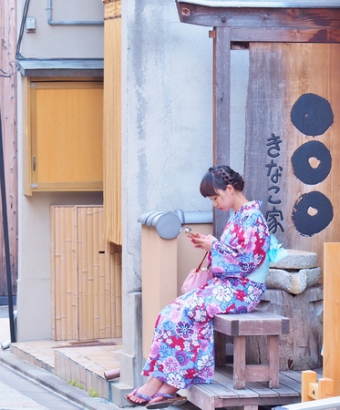 KYOTO, JAPAN - JUNE 08, 2016: Young woman wear a traditional dress called Kimono sitting on wooden bench and playing mobile phone at ancient street in Kyoto, Japan.のeditorial素材