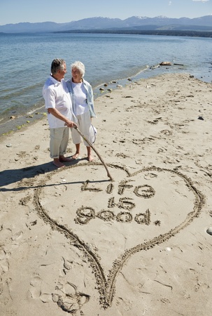 Happy retired couple on beach writing with stick in the sand.の写真素材
