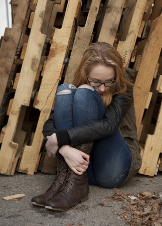 Outdoor photo of young teenage girl seated on ground, hugging knees, looking down at ground.の写真素材