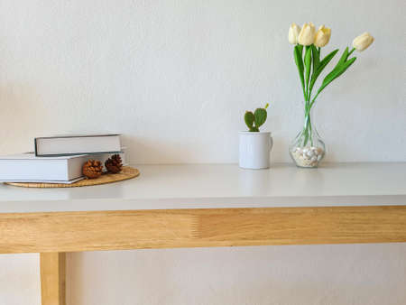 A wooden shelf with a vase of beautiful tulips, cacti and a book on a white cement wall.