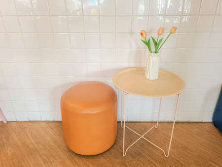 Orange tulips in a modern white vase on a cream colored table beside a round chair. The decoration...