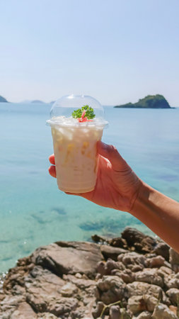 Hand holding glass of fresh milk drink and on stone floor near sea in morning, space for textの写真素材