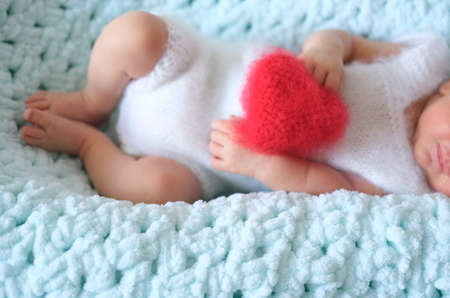 A sleeping newborn boy or girl lies on blue wool. A red heart in her or his hands.Heart shape red...