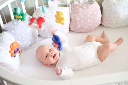 A newborn baby in white clothes lies in the crib and looks at his toys. Baby smiling close upの写真素材