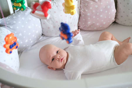A newborn baby in white clothes lies in the crib and looks at his toys. Baby smiling close upの写真素材