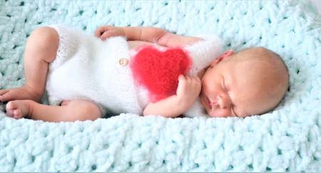 A sleeping newborn boy or girl lies on blue wool. Heart shape red fluffy soft pillow or cushion for Valentines day loveの写真素材