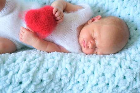 A sleeping newborn boy or girl lies on blue wool. Heart shape red fluffy soft pillow or cushion for Valentines day loveの写真素材