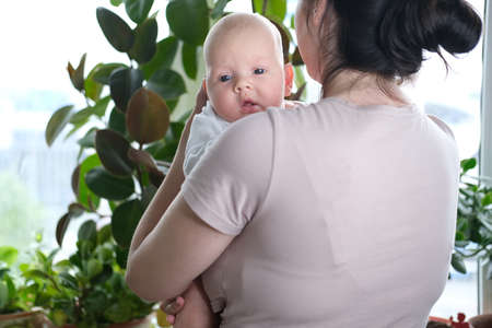 A beautiful young mother holds her newborn son in her arms by the window. Close-up portrait of a childの写真素材