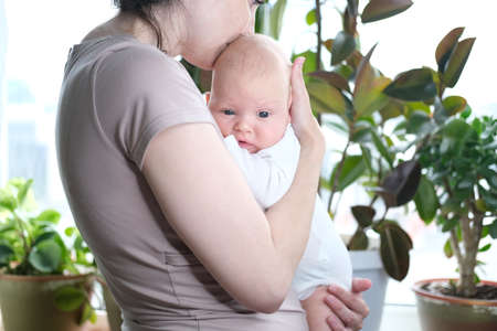 A beautiful young mother holds her newborn son in her arms by the window. Close-up portrait of a childの写真素材