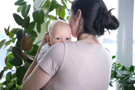 A beautiful young mother holds her newborn son in her arms by the window. Close-up portrait of a childの写真素材