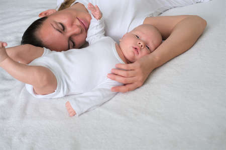 Beautiful young mother lies with her newborn baby on the bed.Portrait of mom and son in white clothes.の写真素材