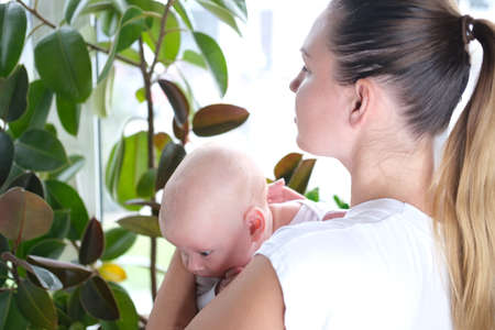 A beautiful young mother holds her newborn son in her arms by the window. Close-up portrait of a child.の写真素材