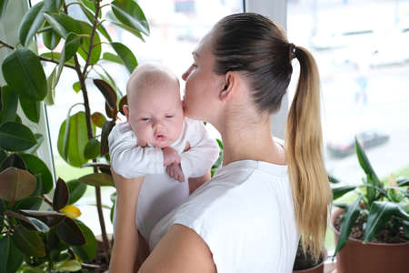 A beautiful young mother holds her newborn son in her arms by the windowの写真素材