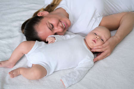 Beautiful young mother lies with her newborn baby on the bed.Portrait of mom and son in white clothes.