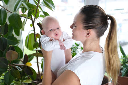 A beautiful young mother holds her newborn son in her arms by the windowの写真素材