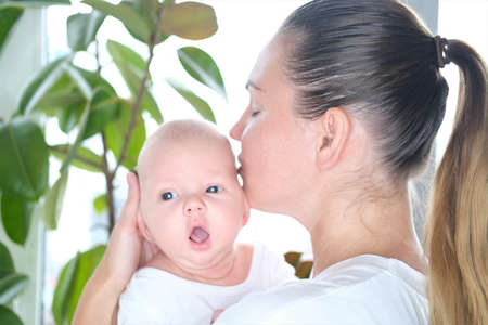 A beautiful young mother holds her newborn son in her arms by the window. Close-up portrait of a child.の写真素材