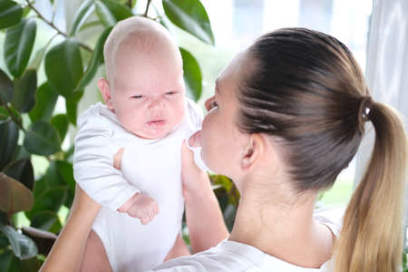 A beautiful young mother holds her newborn son in her arms by the window. Close-up portrait of a child.の写真素材