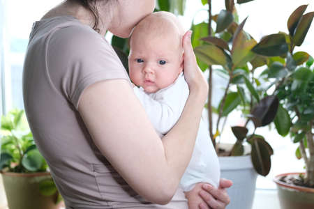 A beautiful young mother holds her newborn son in her arms by the window. Close-up portrait of a childの写真素材