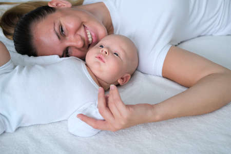 Beautiful young mother lies with her newborn baby on the bed.Portrait of mom and son in white clothes.の写真素材