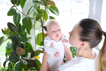 A beautiful young mother holds her newborn son in her arms by the window. Close-up portrait of a child.の写真素材