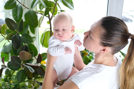 A beautiful young mother holds her newborn son in her arms by the window. Close-up portrait of a child.の写真素材
