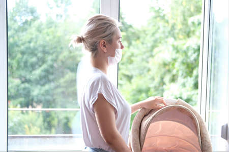 A young mother walks with a newborn baby on the balcony. A girl in a protective mask stands at the window.の写真素材