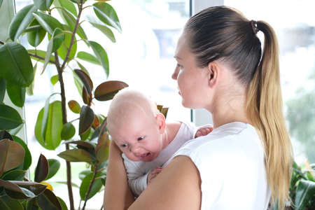 A beautiful young mother holds her newborn son in her arms by the window. Close-up portrait of a child.の写真素材