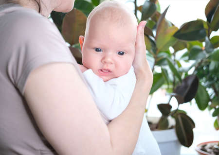A beautiful young mother holds her newborn son in her arms by the window. Close-up portrait of a...