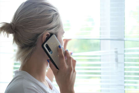 Young beautiful woman in a medical mask is standing by the window and talking on the phone. Prevention of COVID-19.の写真素材