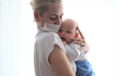 A young woman in a protective mask holds a newborn baby by the window.Portrait of a beautiful young woman and child.の写真素材