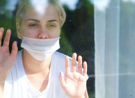 Young beautiful woman in a medical mask stands by the window.Portrait of a girl in a protective maskの写真素材