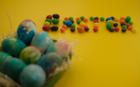 Multicolored Easter eggs lie in a basket on a yellow background, with bright colored sweets,...