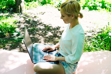 A girl works and communicates with friends on a laptop in the park. The concept is remote work via the Internet and the Covid 19 pandemicの写真素材