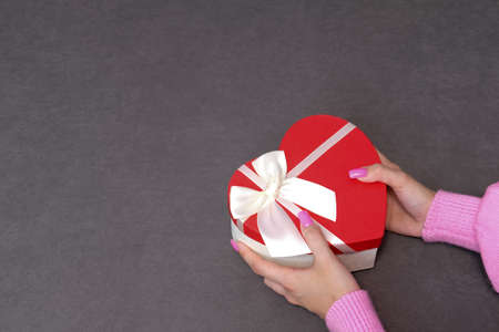 Girl holding a gift in a red box in the shape of a heart. Dark background, selective focus. Concept black friday, holiday, birthday, womens dayの写真素材