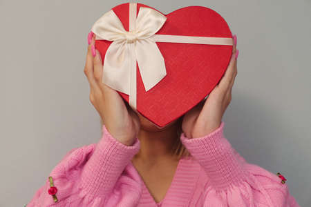 Girl holding a gift in a red box in the shape of a heart.Light background, selective focus.Concept -...